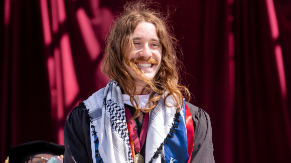 A smiling young man with long red hair wearing commencement regalia.