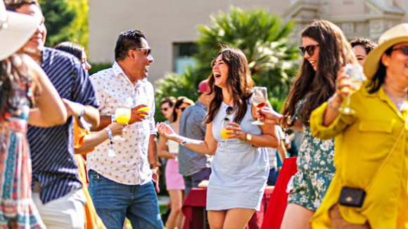 A group of smiling people dancing outside. Several are holding wine or mimosa glasses.
