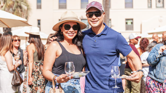 A smiling couple holding wine glasses and plates of food in front of Santa Clara's St. Joseph's Hall.